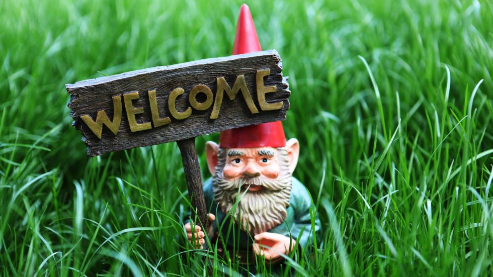 A garden gnome is standing amidst tall grass and is holding a sign in his hand where "Welcome" is written on.