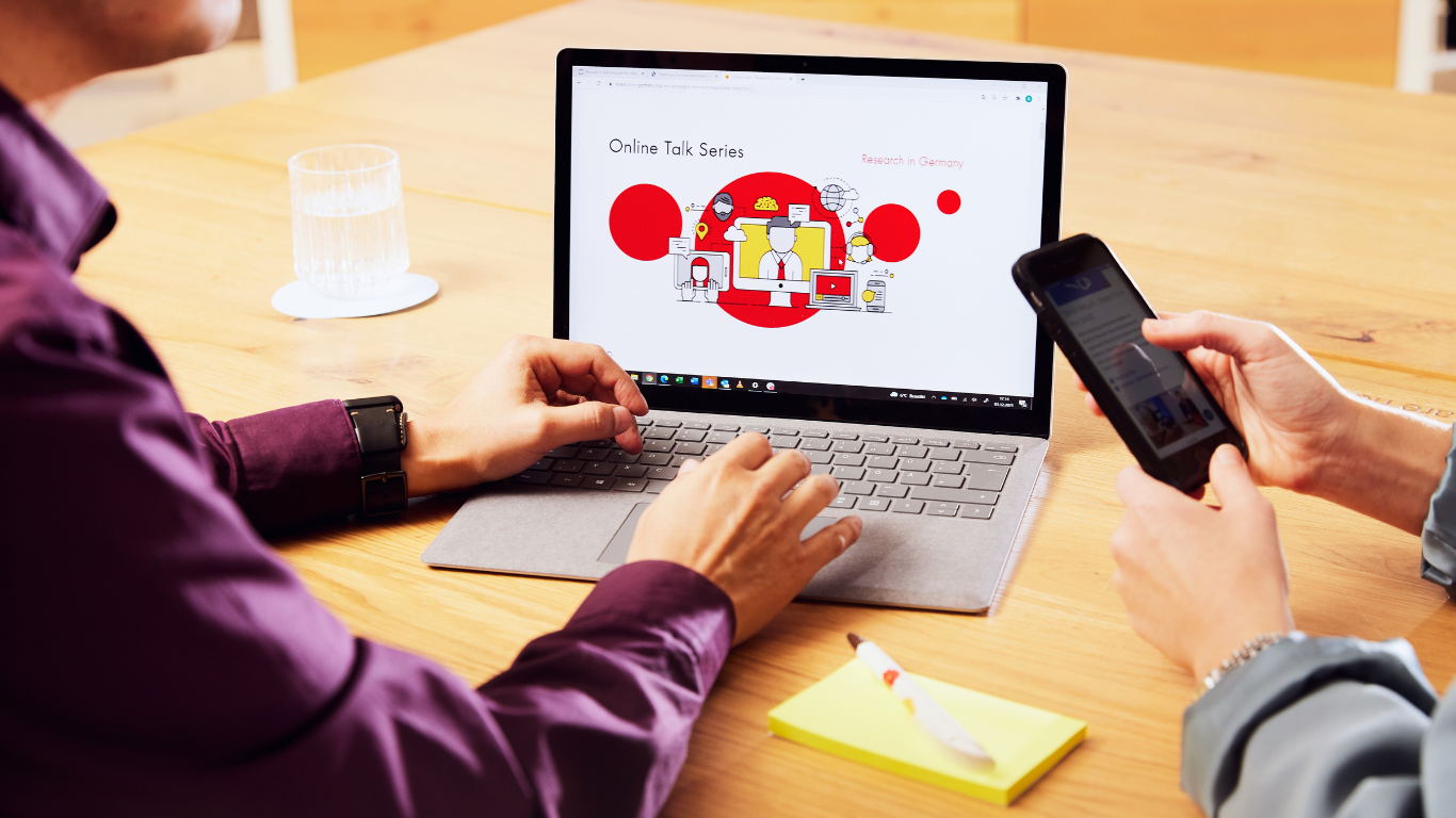 Two people in front of a laptop saying "Online Talk Series", a little notebook and a pen are placed on their table as well. And the one person is using her phone. 