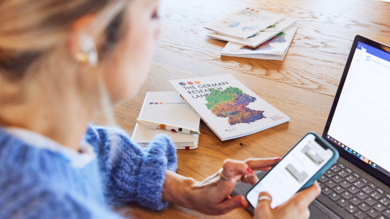 Woman is sitting in front of her laptop, participating in an event and is using her phone as well to register for something online. She is wearing IPods and brochures are placed next to her screen. One can read the title of the top brochure: "The German Research Landscape".