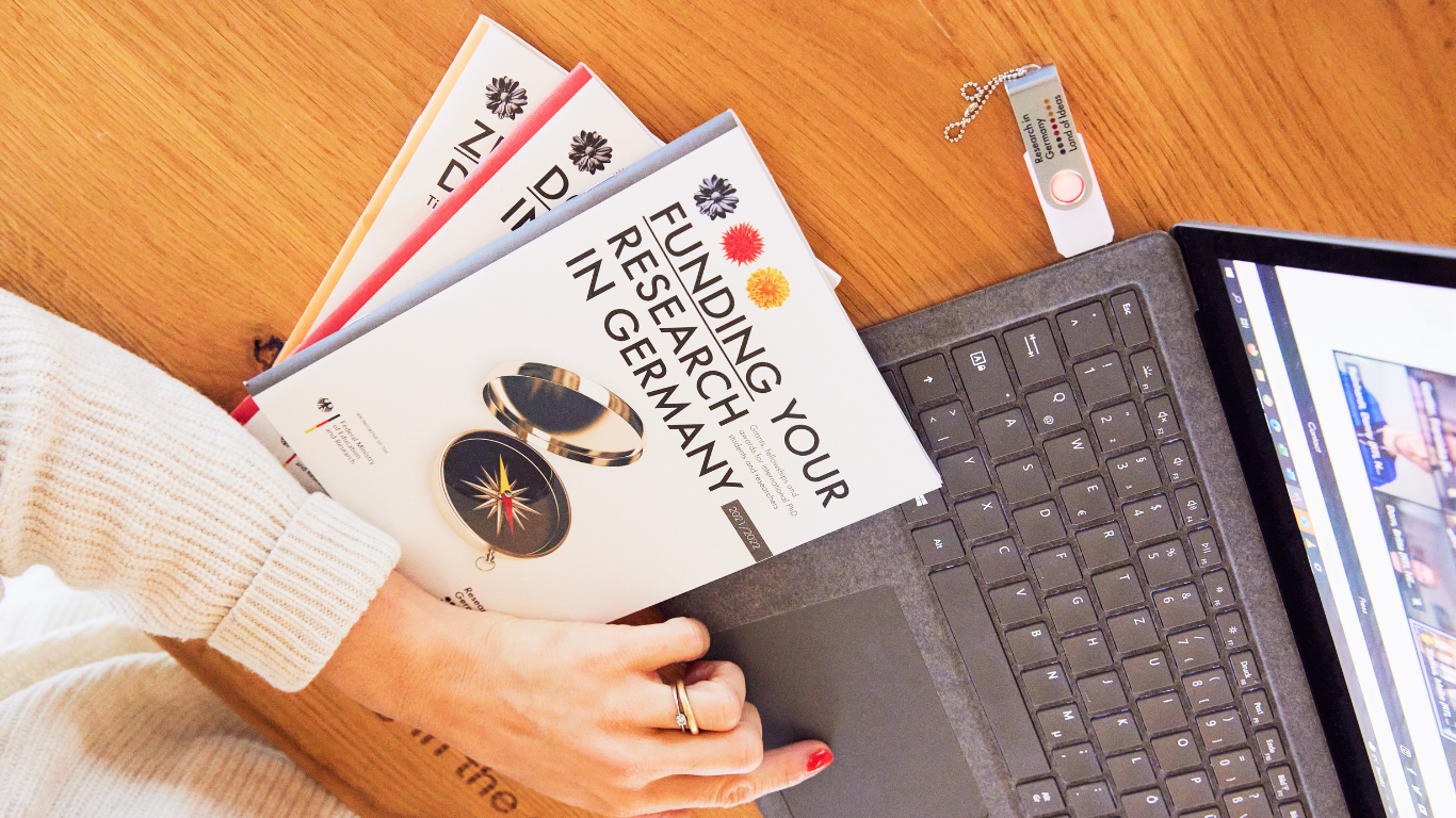 Woman working on her laptop, three brochures are laying next to her, saying "Funding your research in Germany".